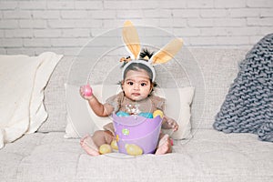 Cute Indian baby girl with pink bunny ears and basket of colorful eggs celebrating Easter holiday