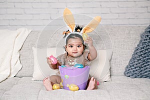 Cute Indian baby girl with pink bunny ears and basket of colorful eggs celebrating Easter holiday