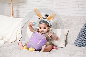 Cute Indian baby girl with pink bunny ears and basket of colorful eggs celebrating Easter holiday