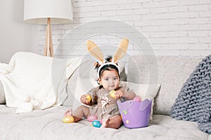 Cute Indian baby girl with pink bunny ears and basket of colorful eggs celebrating Easter holiday