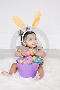 Cute Indian baby girl with pink bunny ears and basket of colorful eggs celebrating Easter holiday