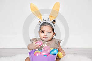 Cute Indian baby girl with pink bunny ears and basket of colorful eggs celebrating Easter holiday