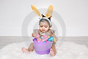 Cute Indian baby girl with pink bunny ears and basket of colorful eggs celebrating Easter holiday