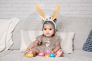 Cute Indian baby girl with pink bunny ears playing with colorful eggs candies toys celebrating Easter holiday