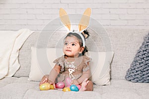 Cute Indian baby girl with pink bunny ears playing with colorful eggs candies toys celebrating Easter holiday