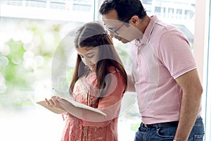 Cute Indian school student girl wears traditional dress near window, holds notebook pencil for doing homework, father teaching