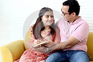 Cute Indian school student girl wears traditional dress sitting with father in living room and doing homework, dad teaching