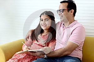 Cute Indian school student girl wears traditional dress sitting with father in living room and doing homework, dad teaching