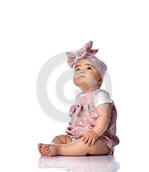 Cute infant baby girl with naive look toddler in polka dot dress and headband with bow sits on the floor looking up