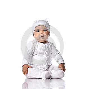 Cute infant baby toddler sits on the floor in white jumpsuit overall and funny hat with ears holding hands at knees