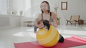 Cute infant boy doing exercises lying on fitness ball while mother holding her legs. Slim fit woman swinging toddler son