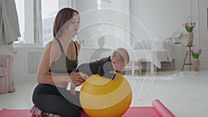 Cute infant boy doing exercises lying on fitness ball while mother holding her legs. Slim fit woman swinging toddler son