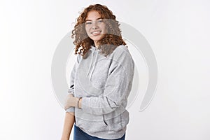 Cute insecure young redhead teenage girl with freckles curly hair wearing grey hoodie smiling touch arm unconfident photo