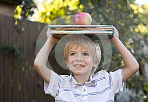 cute joyful 6 year old boy is holding a stack of books and a big apple on his head