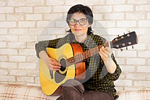 Cute joyful brunette bard girl playing guitar sitting at home on the couch. Copy space. Concept of creativity and music