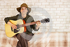 Cute joyful brunette bard girl playing guitar sitting at home on the couch. Copy space. Concept of creativity and music