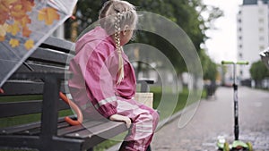 Cute joyful Caucasian girl sitting on bench on rainy day and shaking legs. Side view portrait of little kid in pink