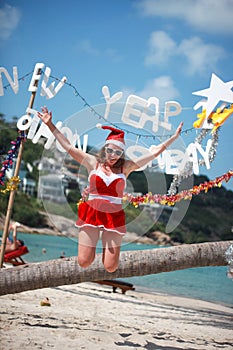 Cute joyful woman jumps in red dress, sunglasses and santa hat on exotic tropical beach. Holiday concept for New Years