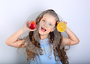 Cute joying grimacing happy kid girl with curly hair style holding citrus orange fruit and red apple in the hands in blue