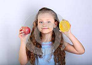 Cute joying smiling kid girl with curly hair style holding citrus orange fruit and red apple in the hands on blue background