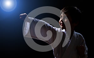 Cute karate boy in white kimono isolated on dark background. Child ready for martial arts fight. Kid fighting at judo training.