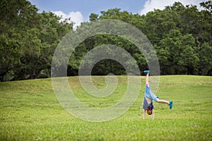 Cute kid boy standing on hands outside on a sunny summer day. Child doing somersault on grass meadow in the park. Active children