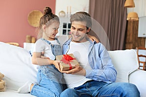 Cute kid daughter make surprise to daddy, little girl presents gift box to father sit on sofa. Father`s day