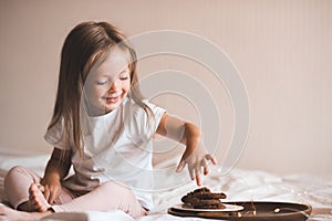 Cute kid girl 3-4 year old wear pajamas sitting in bed eating chocolate pastry on tray with glowing lights close up.