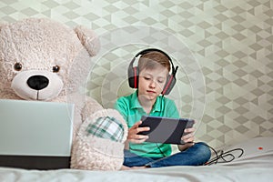 Cute kid in headphones playing on digital tablet next to a toy bear looking in computer at home
