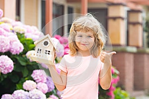 Cute kid playing with small house model outdoors at home garden. Ecology house in childrens hands against backyard.