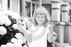 Cute kid playing with small house model outdoors at home garden. Ecology house in childrens hands against backyard.