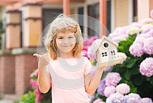 Cute kid playing with small house model outdoors at home garden. Ecology house in childrens hands against backyard.