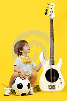 Cute kid sitting on pennyboard with white electric bass guitar, soccer ball and headphone over yellow background.