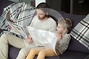 Cute kid son reading newspaper sitting on sofa with dad