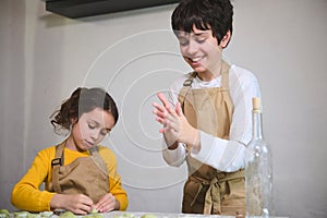 Cute kids in beige chef apron, making dumplings during a cooking class indoors, standing against a white wall background