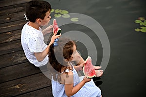 Cute kids with slices of watermelon in their hands, sitting on the pier with their legs lowered into the water and enjoying summer