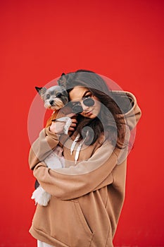 Cute lady in casual clothes stands with biewer terrier dog in hands on red wall background. Vertical