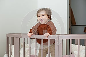 Cute laughing baby standing in round bed. Little girl learns to stand in her crib