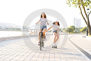 Cute laughing young woman in jeans shorts on bike pulling girl holding on while using skateboard