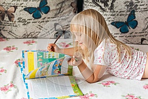 Cute liitle blonde girl lying on a bed and making hometasks in the workbook with a pencil in a hand.