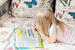 Cute liitle blonde girl lying on a bed and making hometasks in the workbook with a pencil in a hand
