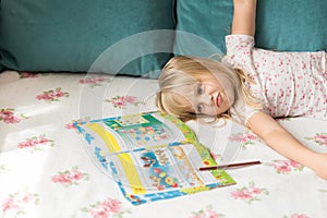 Cute liitle blonde girl lying on a bed and making homework in the workbook with a pencil