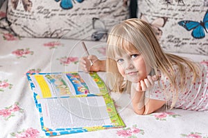 Cute liitle blonde girl lying on a bed and making homework in the workbook with a pencil.