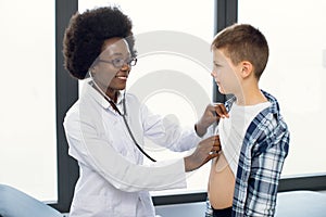 Cute likable Caucasian teen boy and joyful African woman doctor, during doctors checkup. Pediatrist examinates young