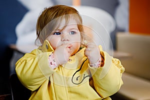 Cute little adorable baby girl sitting in indoor cafe. Happy toddler child eating bread. Colorful baby fashion clothes