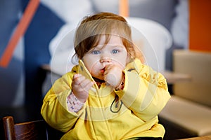 Cute little adorable baby girl sitting in indoor cafe. Happy toddler child eating bread. Colorful baby fashion clothes