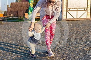 Cute little adorable blond toddler boy making first steps with mother support at city park at evening sunset time. Happy funny