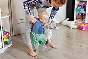 Cute little adorable blond toddler boy making first steps with mother support in playroom at home. Happy funny child