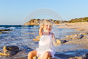 A cute little adorable toddler girl in white clothes playing with sand and shells on the empty beach on a warm sunny summer day. H