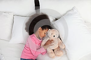Cute little African-American girl with teddy bear sleeping in bed